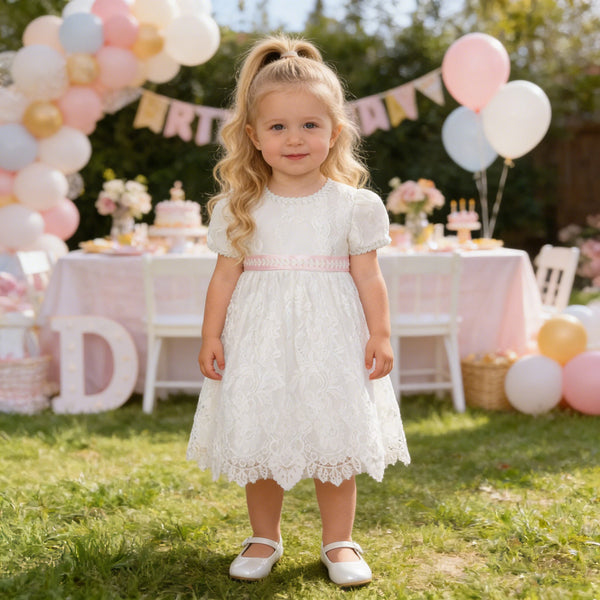 Toddler girl in knee-length white lace baptism dress with pink waist sash, embroidered short sleeves, round neckline
