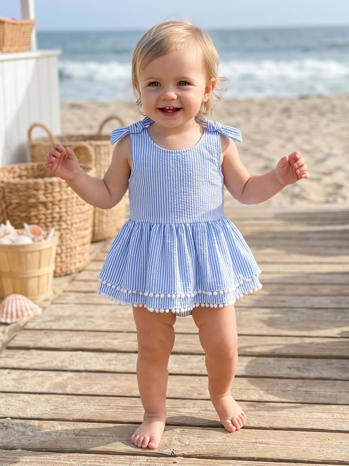 Baby girl wearing blue and white seersucker striped romper with frilly hem and pom-pom trim at the beach.
