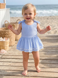 Baby girl wearing blue and white seersucker striped romper with frilly hem and pom-pom trim at the beach.