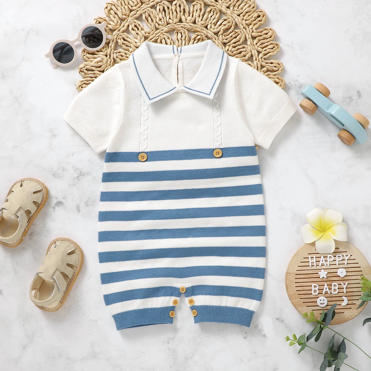 A baby knit romper laid flat on a white marble surface with styled props including a woven mat, sunglasses, sandals, a wooden toy car, a flower, and a small sign. The romper is off-white with blue horizontal stripes across the lower half and a blue collar, accented by small gold-tone buttons.