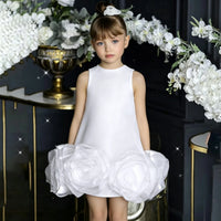 A young girl with a high bun and blue eyes wearing a sleeveless white dress with a large ruffled white fabric flower at the hem, standing in a dark interior decorated with white flowers and gold accents.
