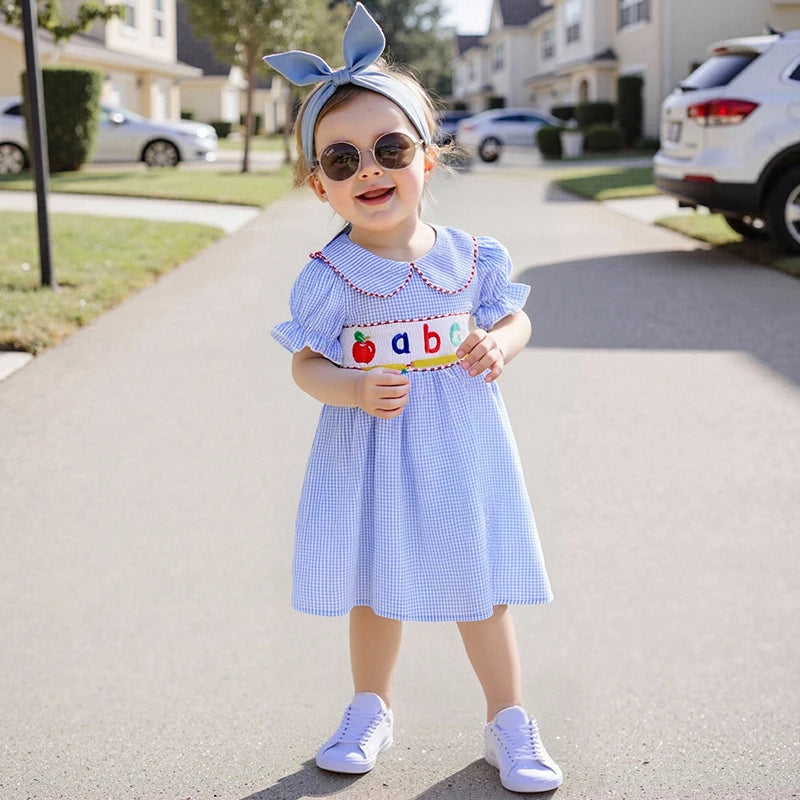 A stylish toddler wearing the ABCs Embroidered Girls Dress - Peter Pan Collar with a headband and sunglasses, smiling while standing on a sunny street.
