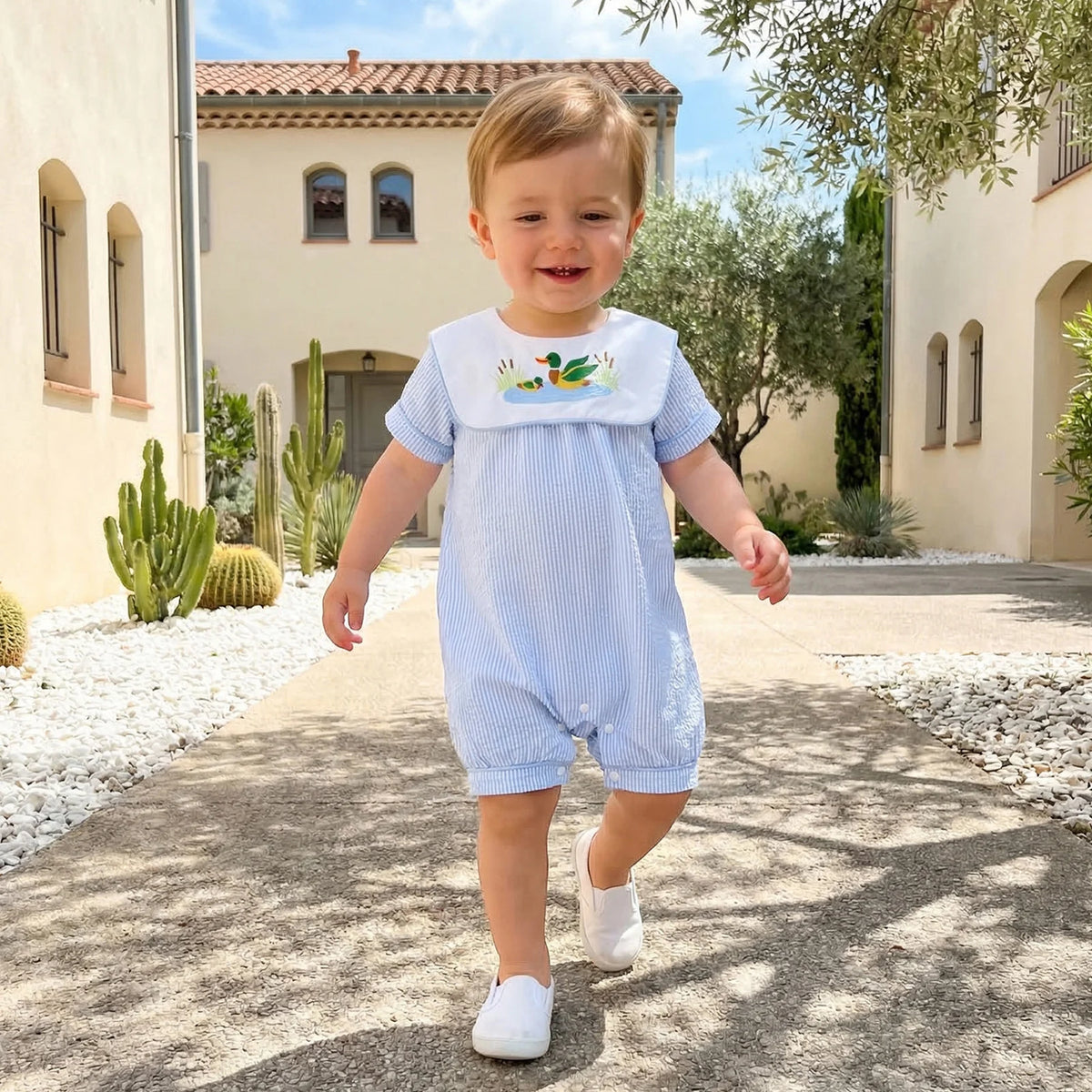 Toddler in Boca Blue bibbed seersucker romper with mallard duck embroidery, walking outdoors.