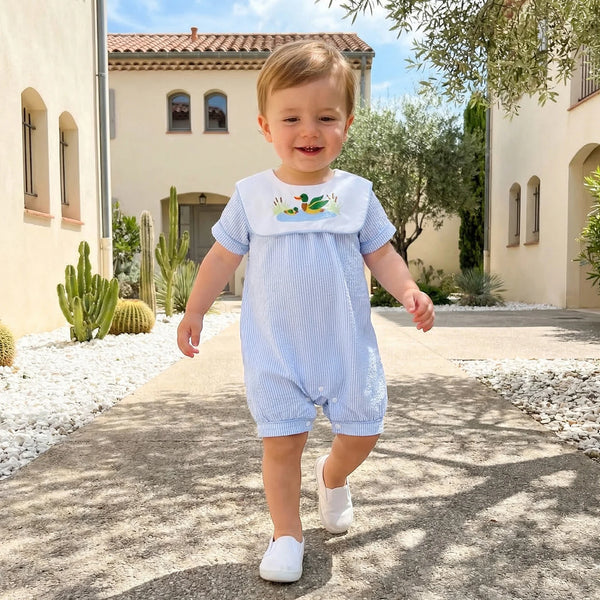 Toddler in Boca Blue bibbed seersucker romper with mallard duck embroidery, walking outdoors.