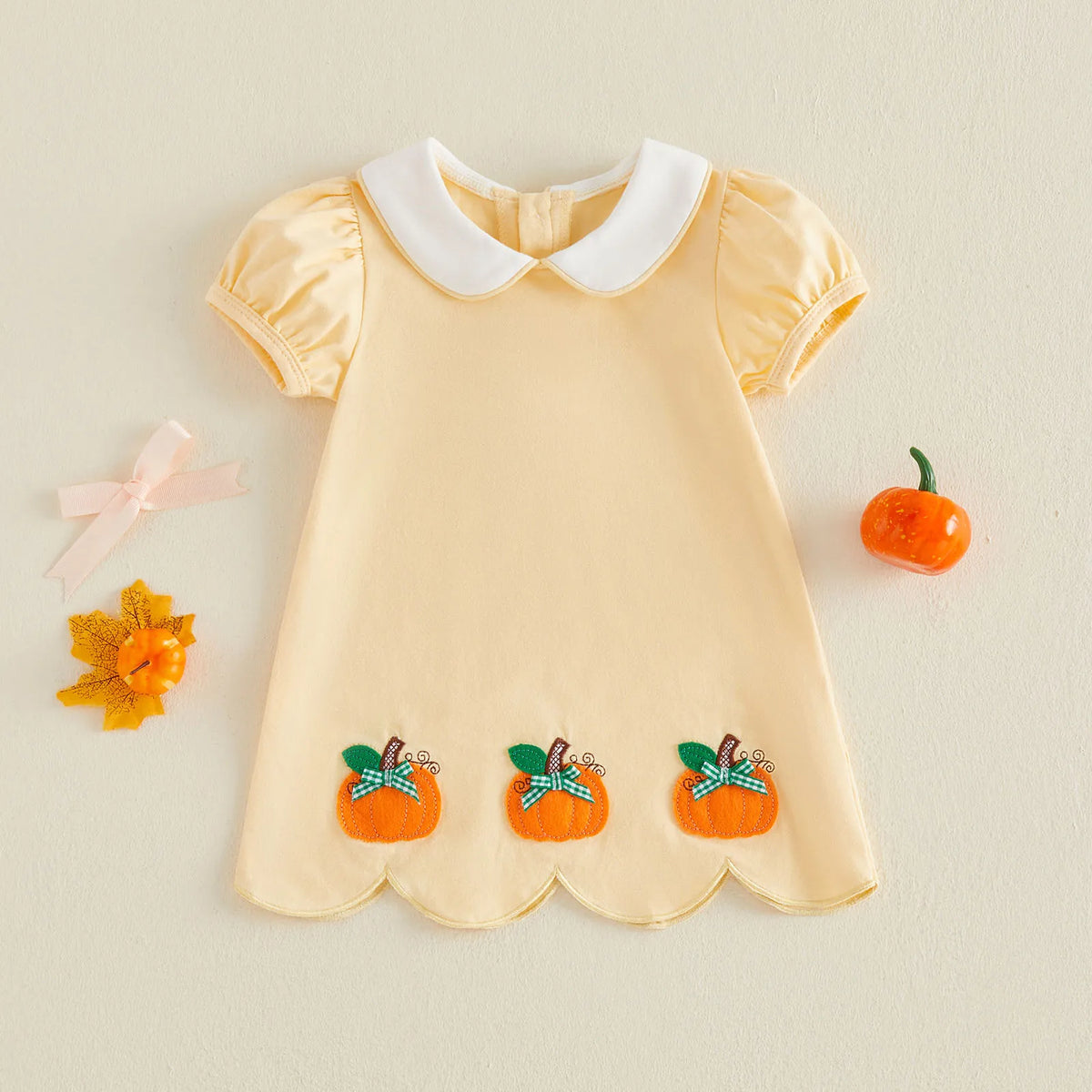 A pale yellow baby dress with a white Peter Pan collar, short puff sleeves, and three embroidered orange pumpkin motifs with green leaves and blue bows near the hem. The dress is laid flat on a light beige background with small decorative props including a pink bow, a yellow flower, and a small orange pumpkin.