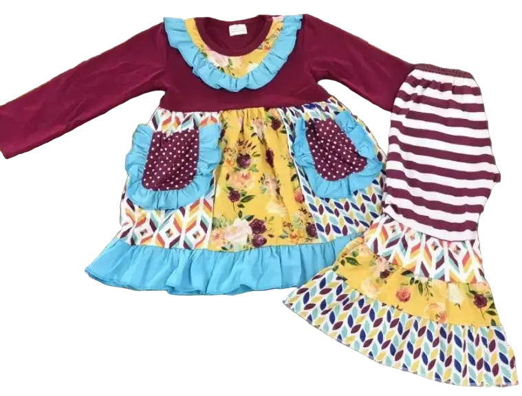 A two-piece toddler girl's outfit laid flat on a wooden floor, featuring a long-sleeve dress with a burgundy top, light blue ruffle trim, and a yellow floral skirt with patterned panels, paired with matching striped leggings.