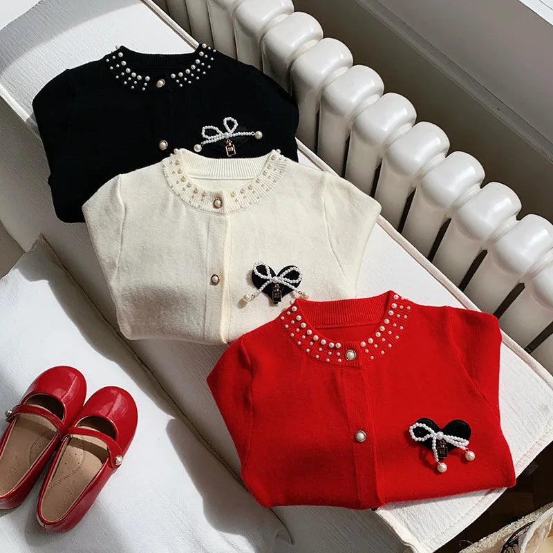 Bows and Pearls Little Girls Cardigan laid out with red, cream, and black options alongside cute red shoes, emphasizing the elegant style perfect for young girls.