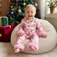 Baby girl wearing pink satin Christmas pajama set with Santa print and fuzzy cuffs, sitting before a decorated tree with gifts.