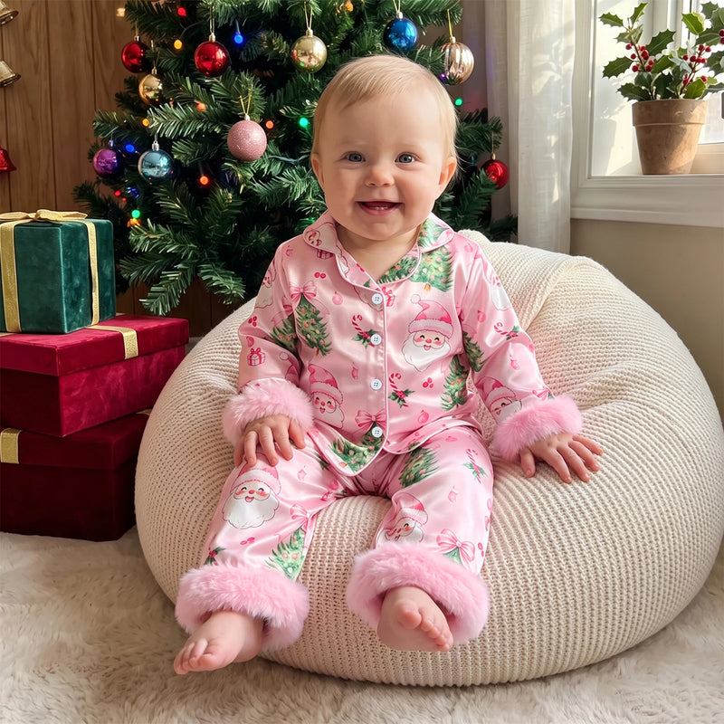 Baby girl wearing pink satin Christmas pajama set with Santa print and fuzzy cuffs, sitting before a decorated tree with gifts.