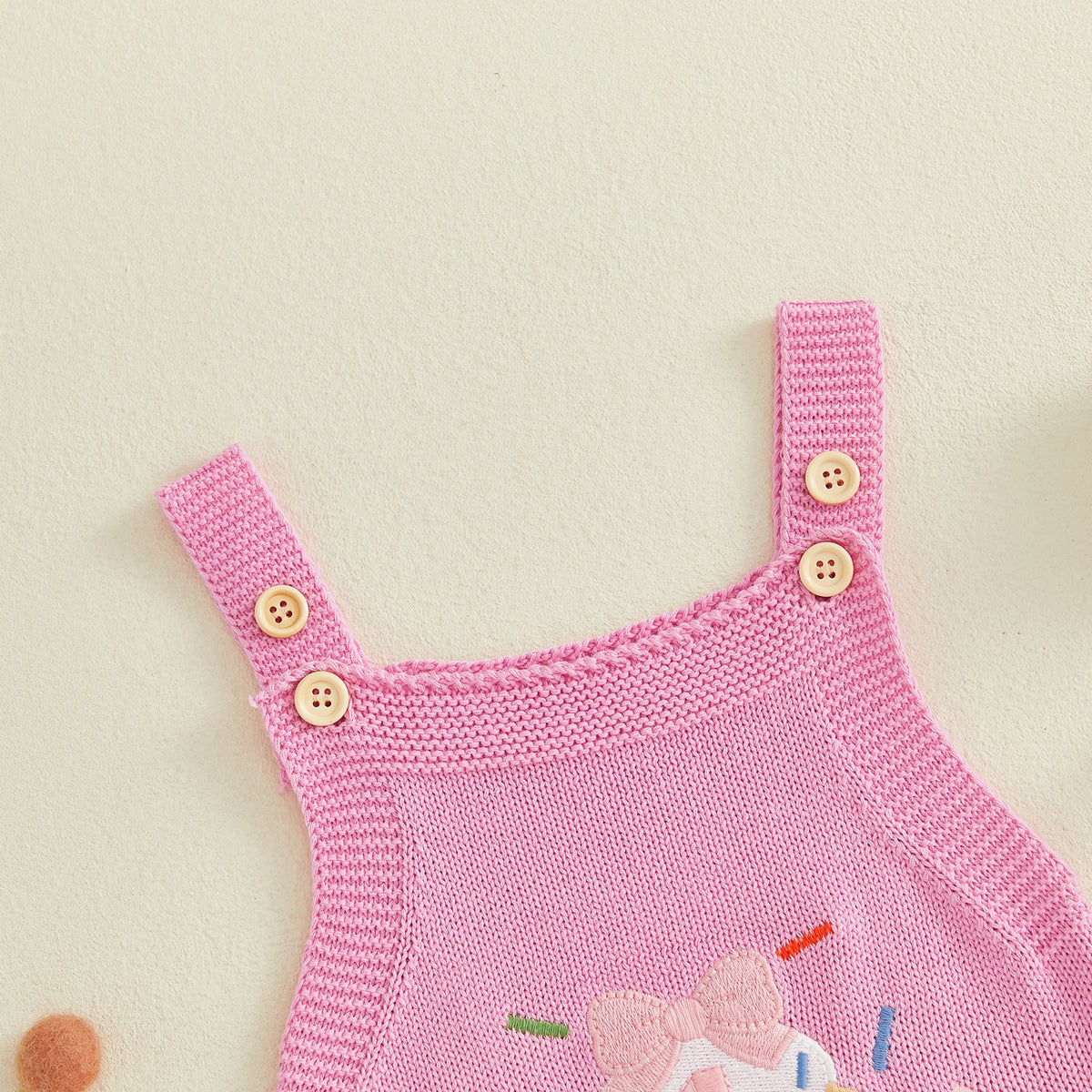 Pink knitted romper overalls with shoulder straps, cream buttons, and cupcake embroidery for a first birthday outfit