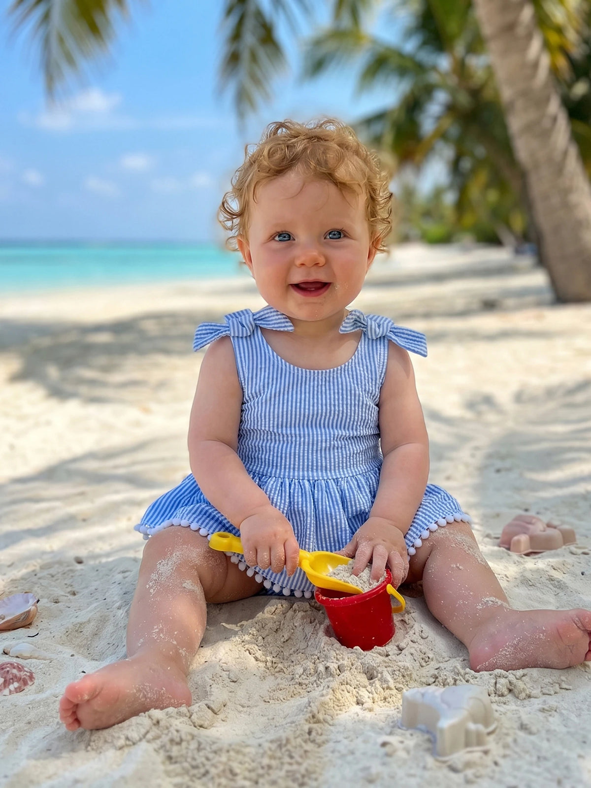 Baby in blue seersucker stripe frilly romper with shoulder bows at the beach.