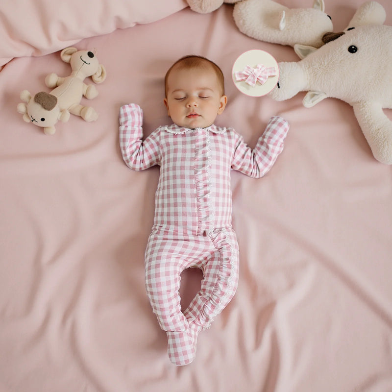 Comfortable baby girl resting in the Checks and Frills Footie Set in Poinciana pink, surrounded by stuffed toys and a cozy blanket.