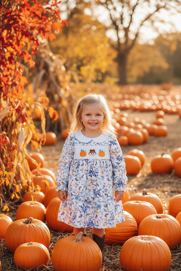 A long-sleeve baby dress with a blue and yellow floral print on a white background, featuring a smocked chest panel with embroidered pumpkins and a cowboy hat.