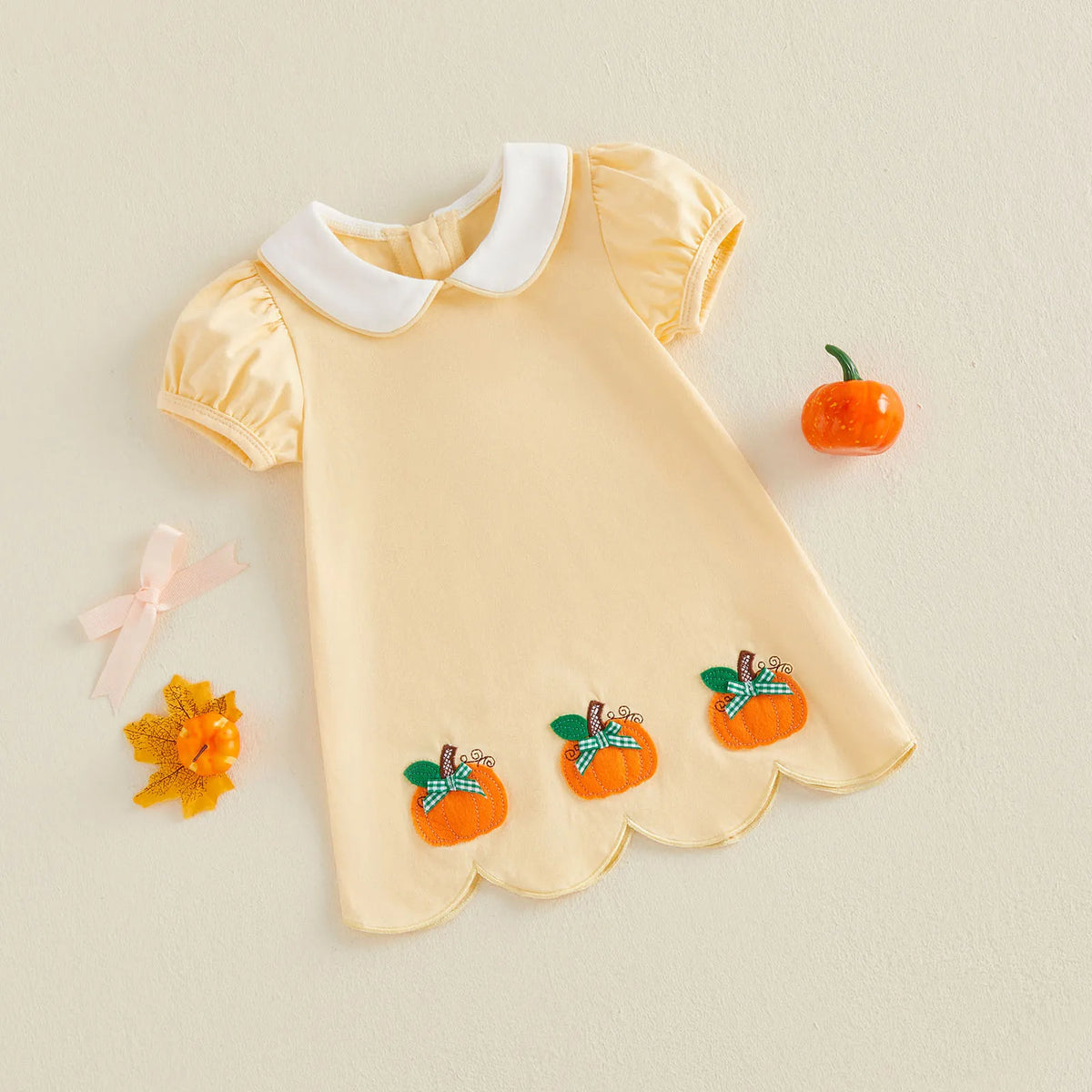 A baby girl's short-sleeve dress in pale yellow with a white Peter Pan collar, decorated with three orange pumpkin appliques on the hem. The dress is styled flat-lay on a textured beige background with autumn-themed props including a small orange pumpkin, a pink bow, and a yellow leaf.