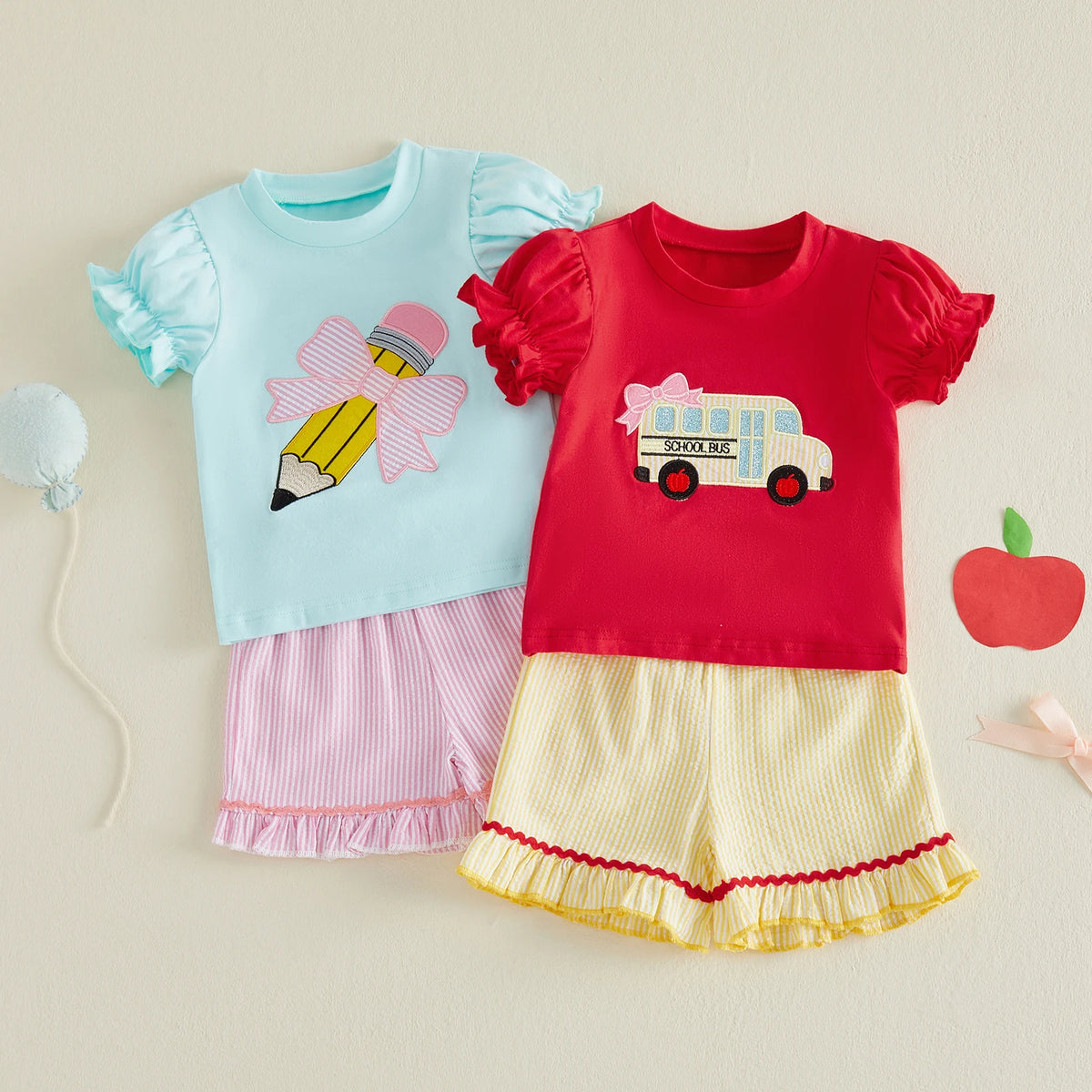 Two toddler back-to-school outfits: blue top with pencil bow and pink shorts; red top with bus patch and yellow shorts.