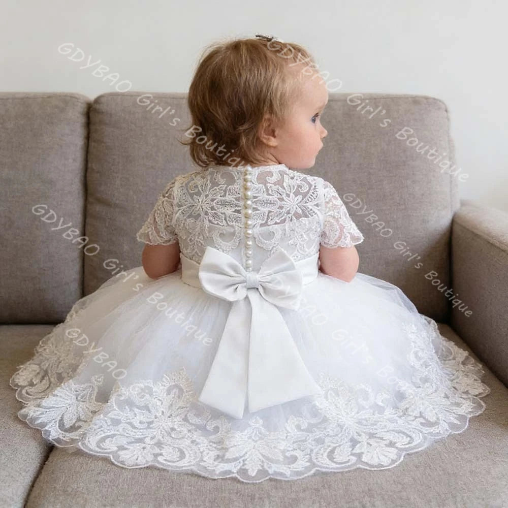 White lace flower girl dress featuring a tiered tulle skirt, applique hem, faux pearl back buttons, and a large oversized satin bow.