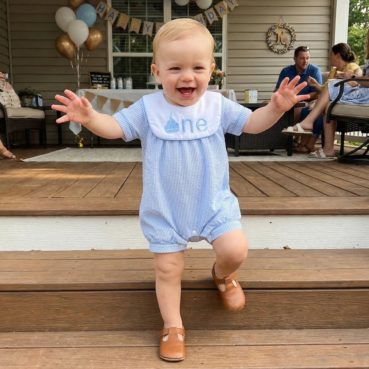Baby in a blue outfit with 'one' bib on a wooden deck, celebrating a birthday.
