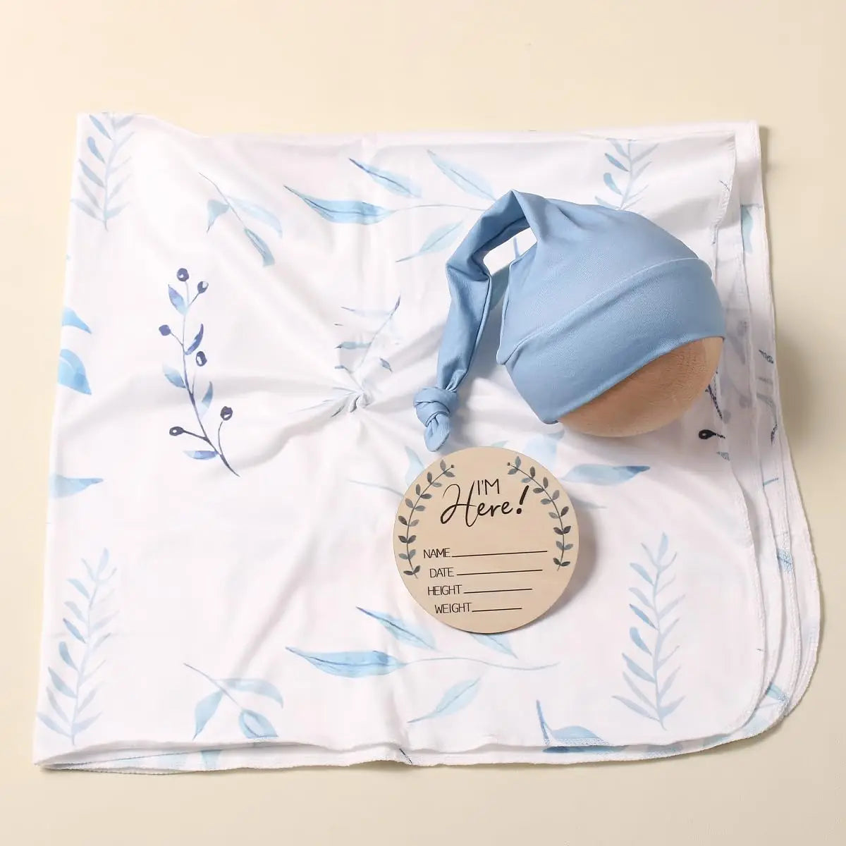 White swaddle blanket with blue leaf pattern, matching blue elf hat, and "I'm Here" birth announcement tag set for newborns.