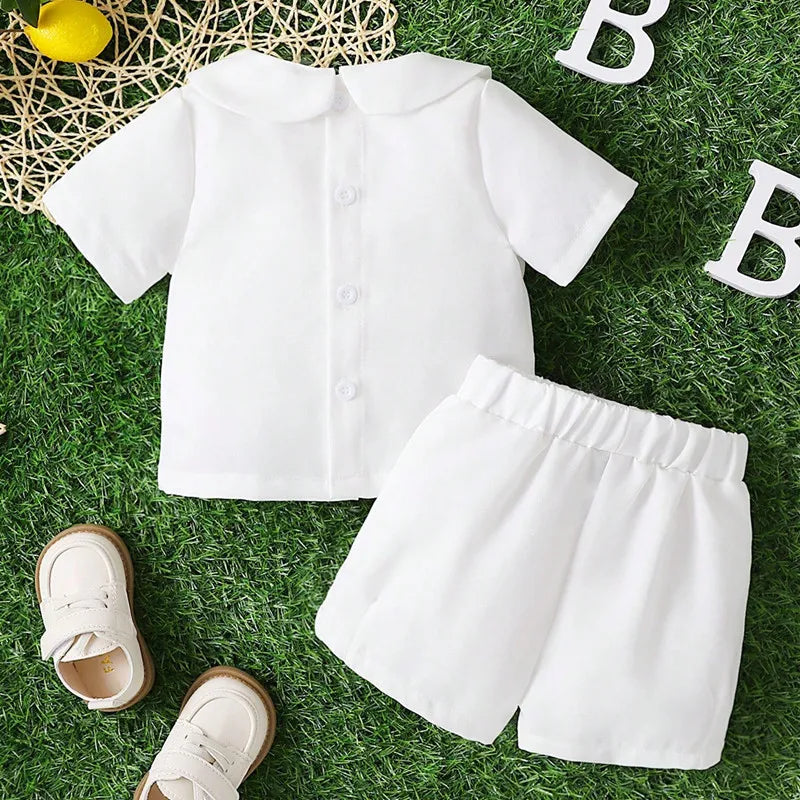 White boys 2-piece baptism set with Peter Pan collar shirt and shorts on grass, paired with white shoes for warm weather.
