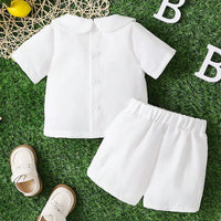 White boys 2-piece baptism set with Peter Pan collar shirt and shorts on grass, paired with white shoes for warm weather.