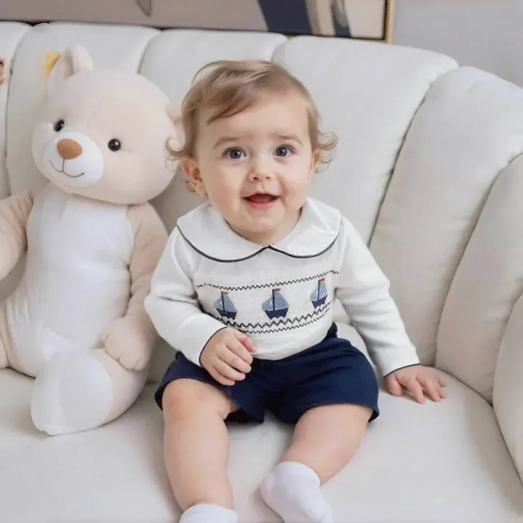 toddler boy wearing nautical toddler outfit with white top and navy shorts sitting on white couch next to teddy bear