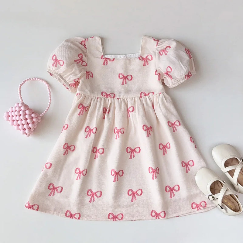 Sweet Little Bows party dress featuring a charming pastel pink fabric with adorable pink bows, perfect for girls aged 3 to 7. The dress includes puff sleeves, a square neckline, and a flared skirt, ideal for twirling at any special occasion.