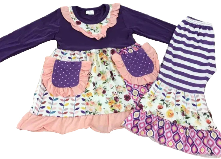 A children's two-piece outfit laid flat on a wood floor, featuring a purple long-sleeve top with a floral and ruffled collar, a matching floral skirt with purple polka-dot pockets and pink ruffles, and a separate matching top piece with purple and white horizontal stripes.
