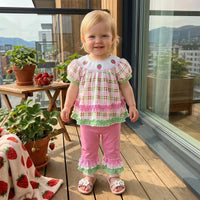 Toddler girl in a pink-green plaid frilly two-piece outfit with strawberry embroidery, standing on a balcony.