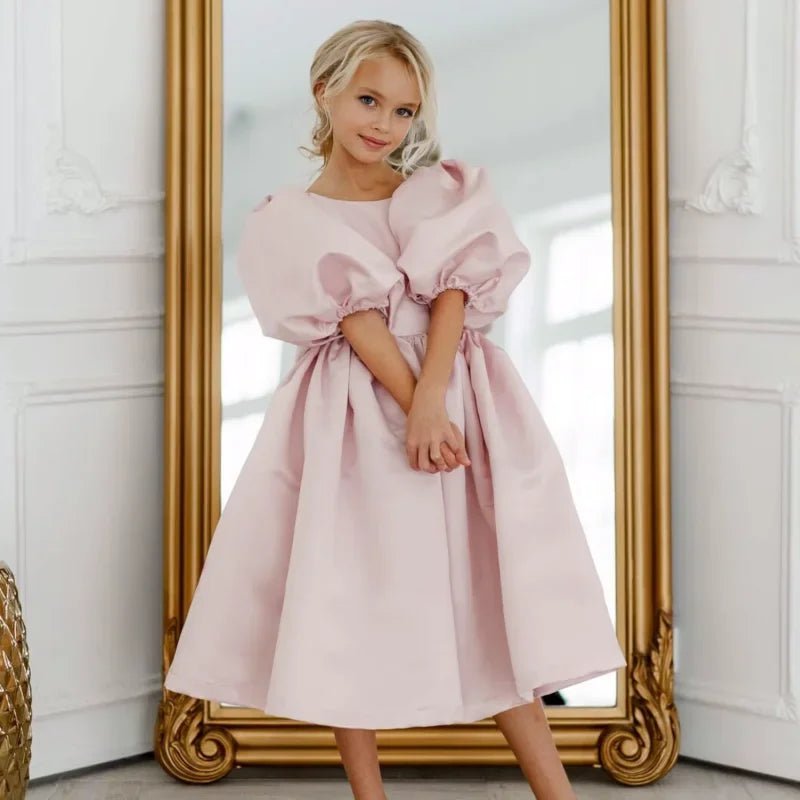 A young girl wearing the 'Arora' Puffed Sleeved Party Dress in princess pink, showcasing dramatic puffed sleeves and a scooped neckline in front of a mirror.