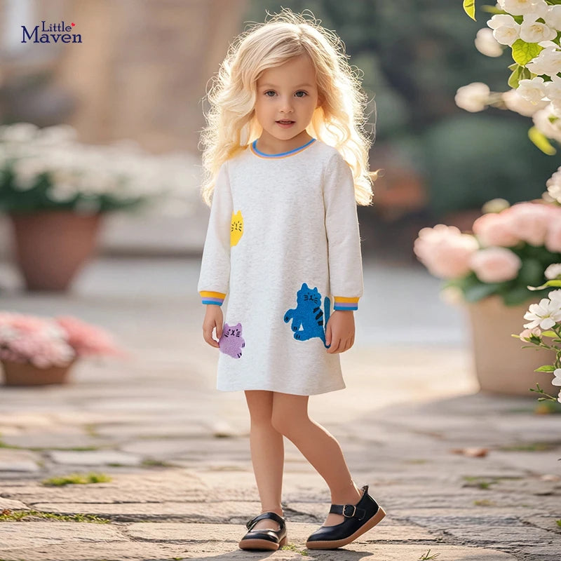 A young girl wearing the 'Here, Kitty Kitty!' Adorable Textured Appliqued Dress featuring colorful cat appliqués, smiling while standing outdoors in a playful pose.