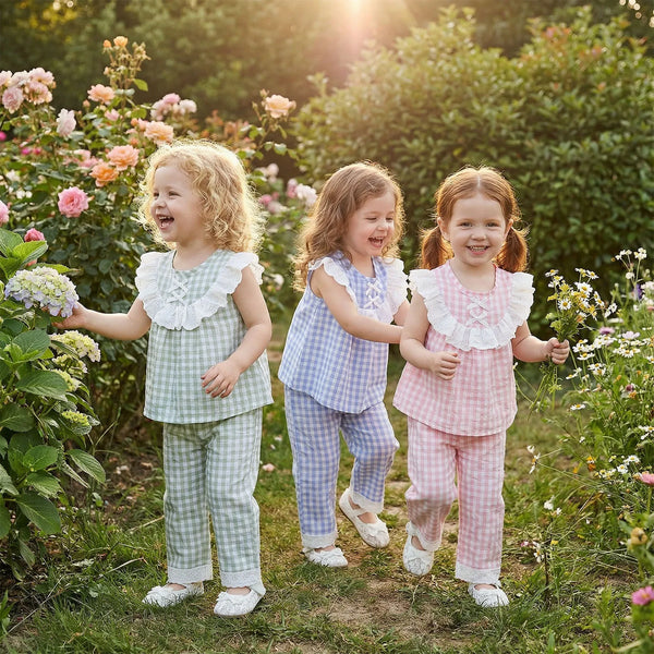 Three little girls in pastel gingham two-piece pant sets in pink, blue, and green among garden blooms.