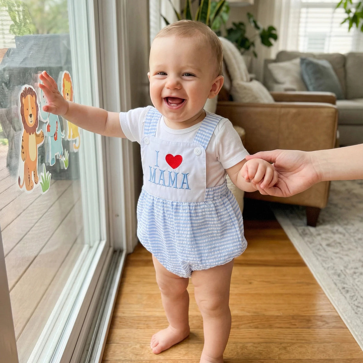 Smiling baby in blue-and-white check bubble romper embroidered I Love Mama by a window.