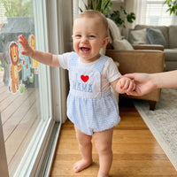 Smiling baby in blue-and-white check bubble romper embroidered I Love Mama by a window.
