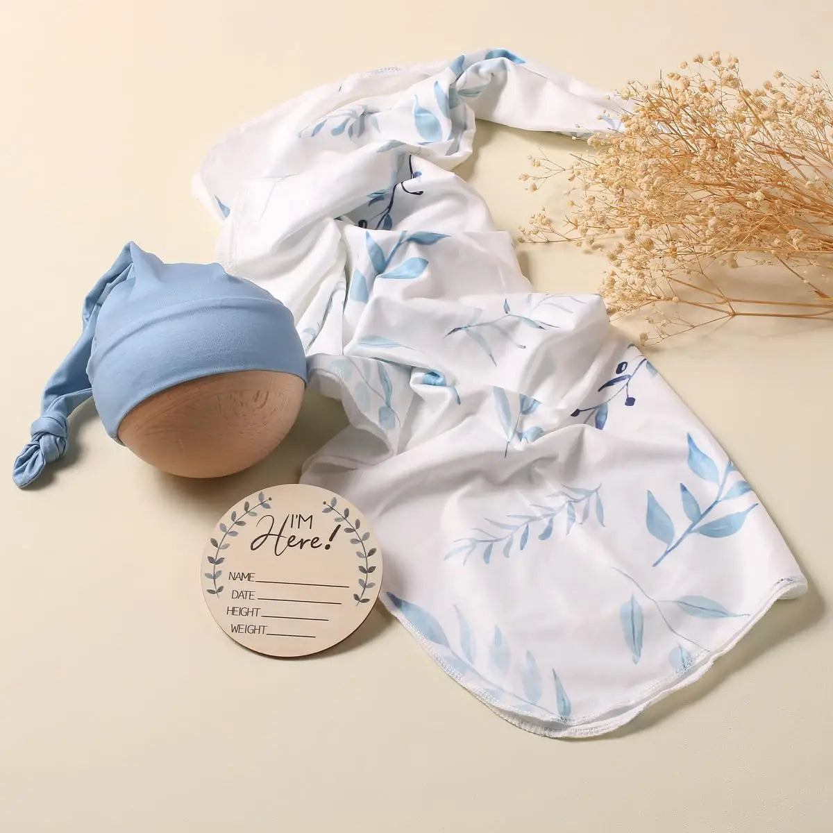Blue leaf-print baby swaddle and matching cap set with wooden birth record tag and dried flowers.