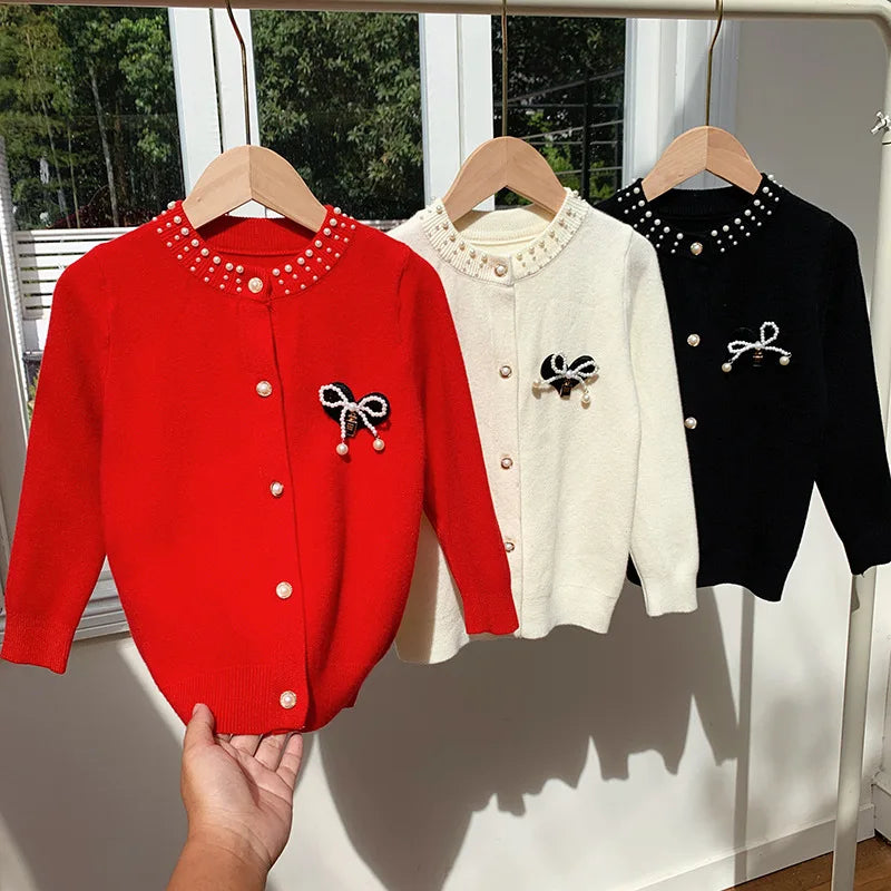 Bows and Pearls Little Girls Cardigan displayed in three classic colors: Palm Way Red, Soft Apricot Cream, and Chic Black, showcasing the charming design with faux pearl buttons and bow accent.