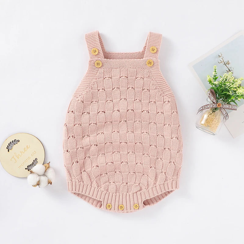 A light pink knit baby romper with gold buttons, laid flat on a white background with decorative props including cotton bolls, a round wooden tag, and a small glass vase with greenery.