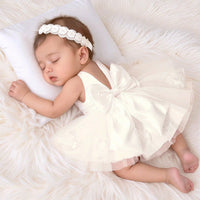 Newborn sleeping girl in a white two-piece special-occasion dress with an oversized back bow and beaded headband.
