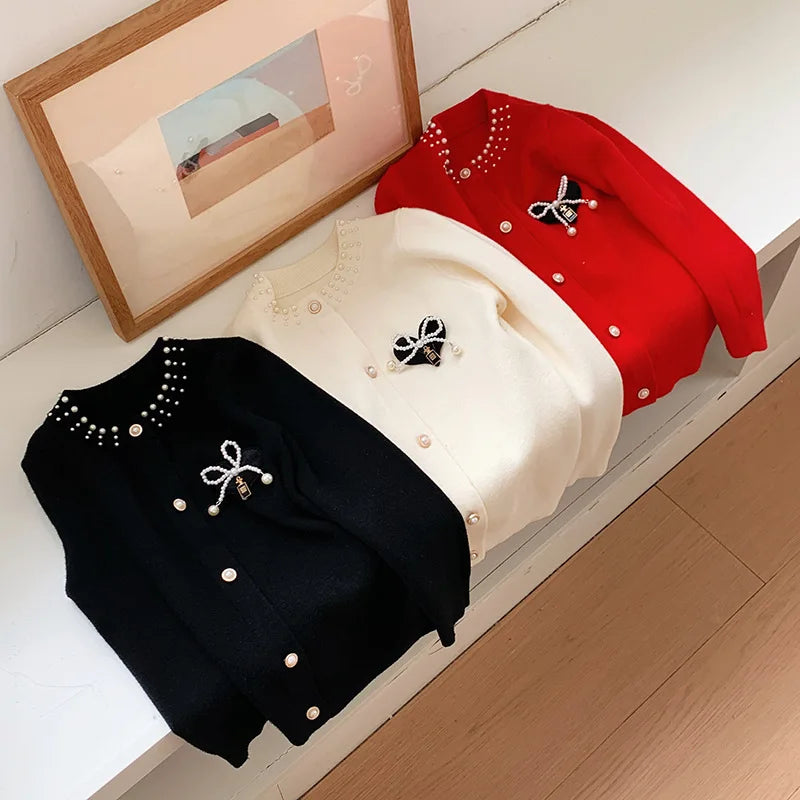 Three Bows and Pearls Little Girls Cardigans in red, cream, and black elegantly arranged next to a framing picture, highlighting their charming designs.