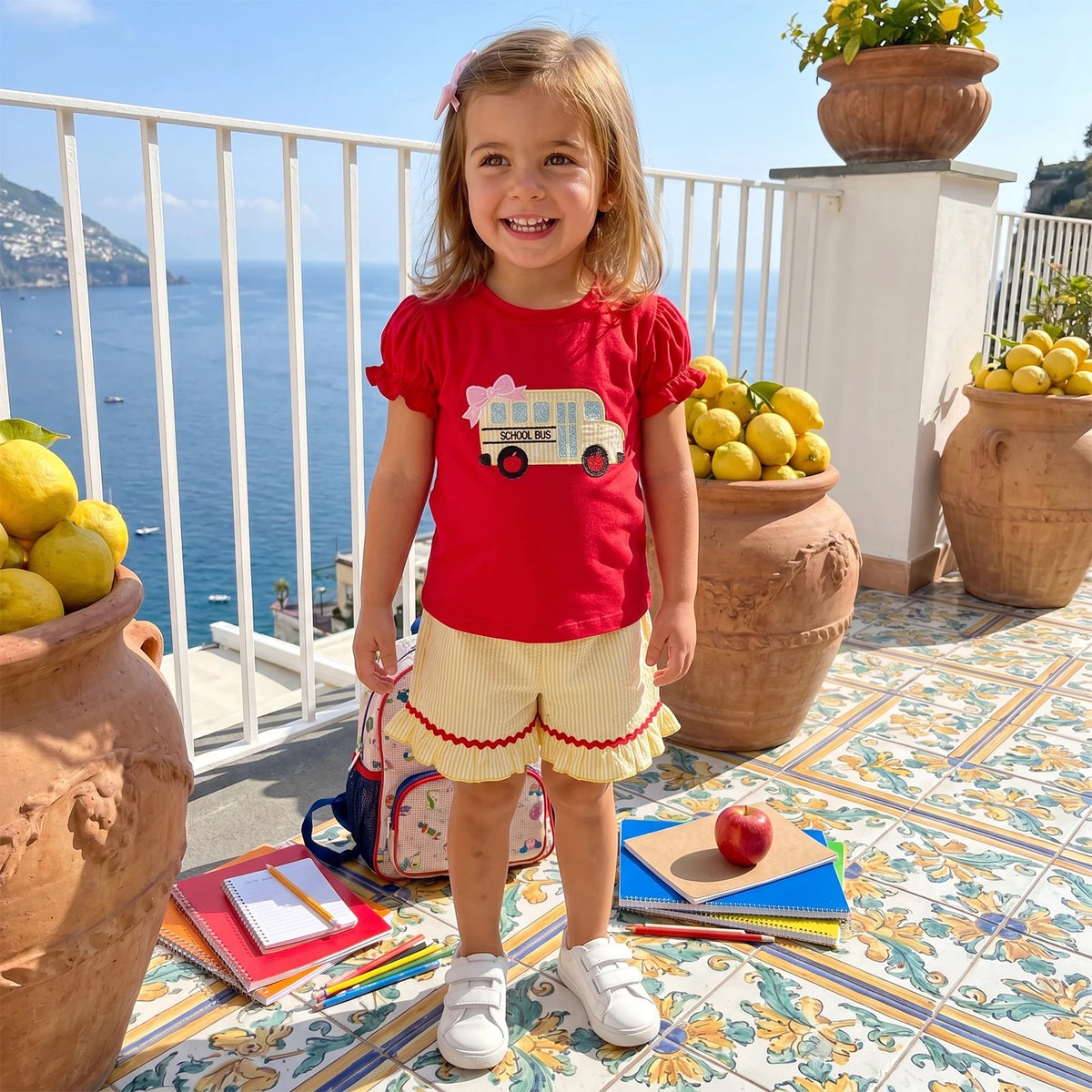 Toddler girl wearing red school-bus embroidered top and striped yellow seersucker shorts with frilly trim.