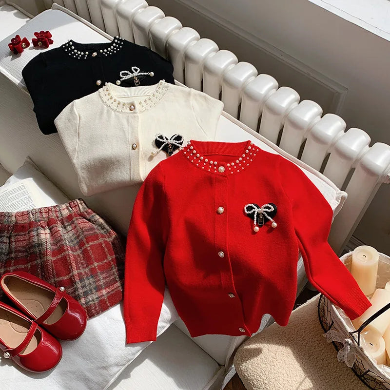 The Bows and Pearls Little Girls Cardigan in classic red, cream, and black, paired beautifully with a plaid skirt and red shoes, ideal for stylish occasions.