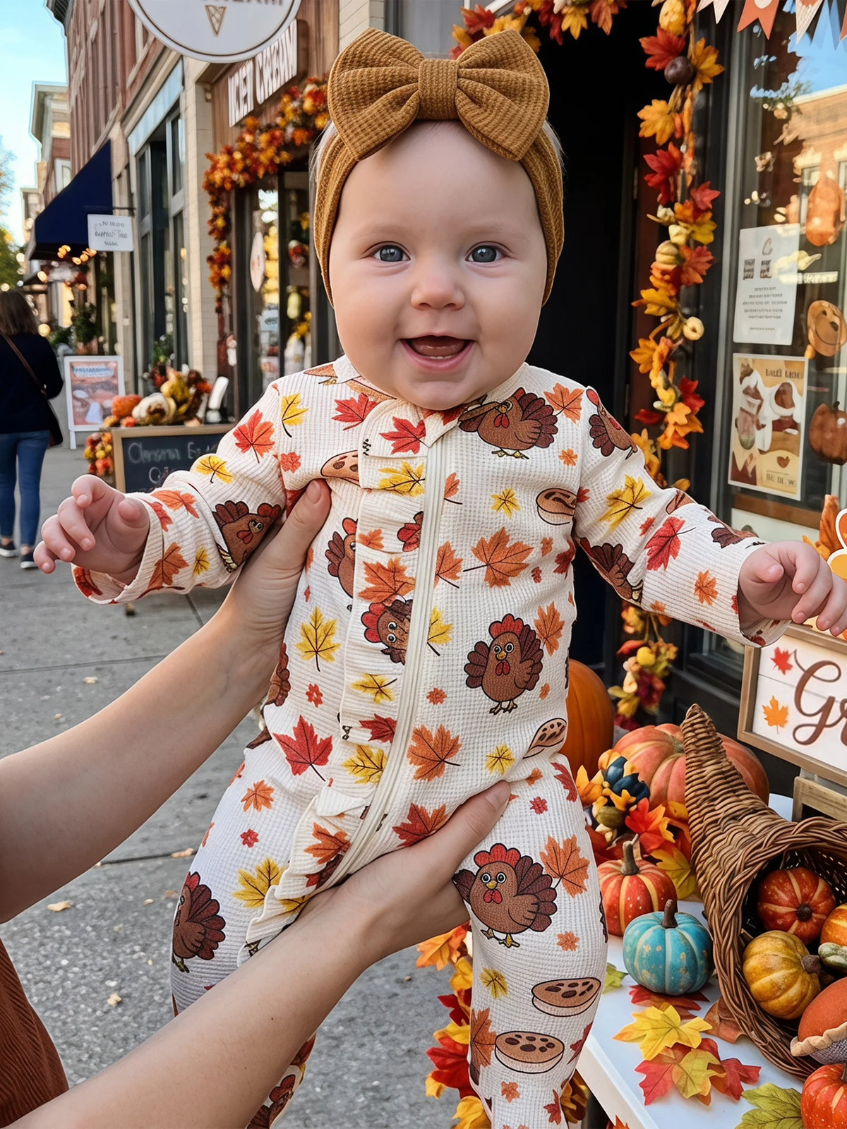 Baby wearing a turkey and leaf print footed romper with front zipper and mustard headbow, held against fall decorations.