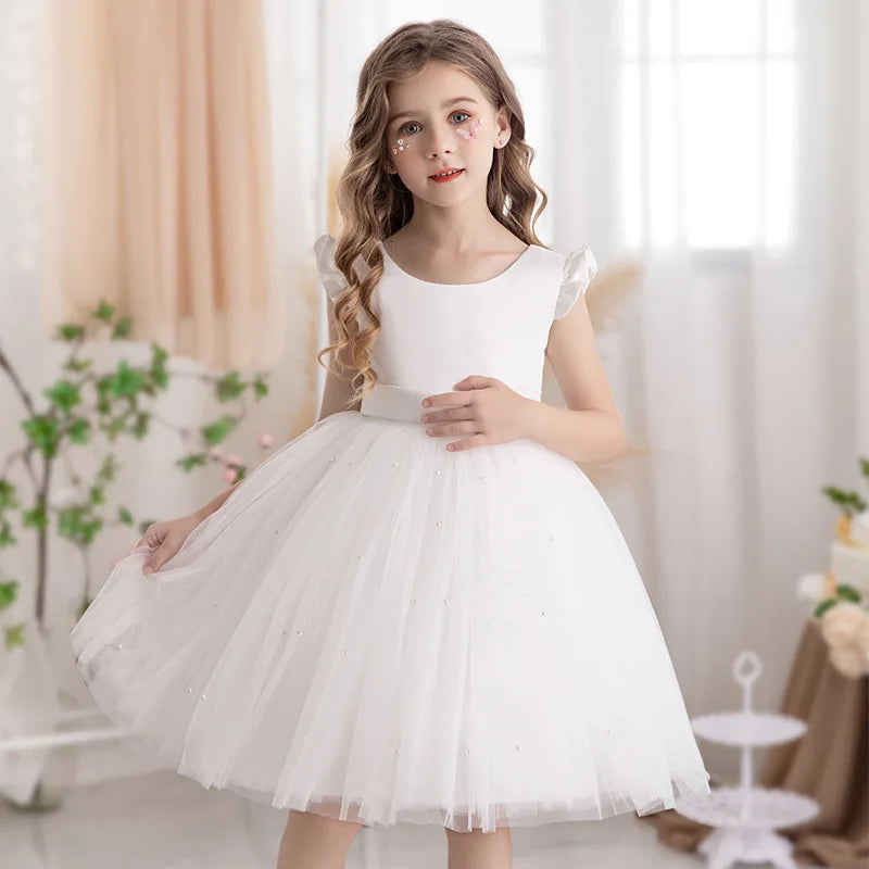 A girl wearing a white 'Chantelle-Elise' V-Back Occasion Dress, featuring a fluffy tulle skirt and a cute bow, perfect for special occasions.