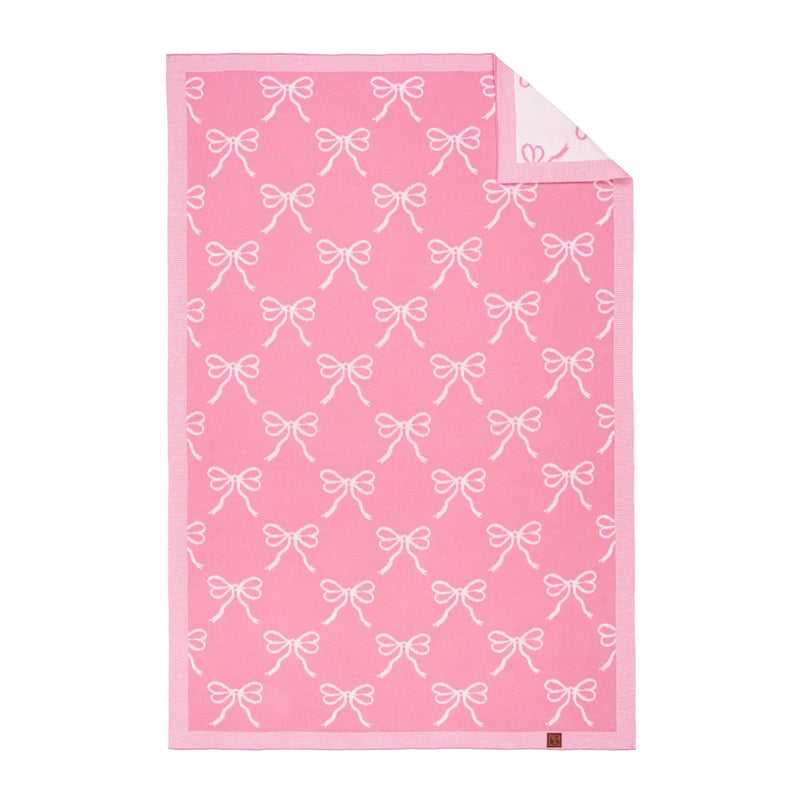 Pink jacquard baby blanket with white bow pattern, folded corner revealing cream reverse side and brown brand tag