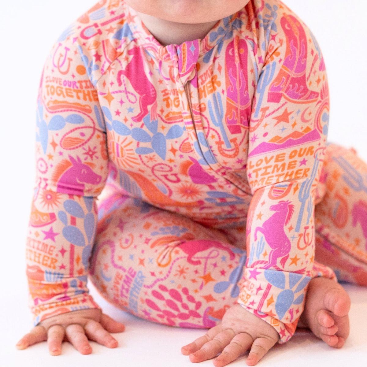 Baby in The Second-Skin Convertible Romper Saddle Up; vibrant pink, orange, blue pattern with zipper front.