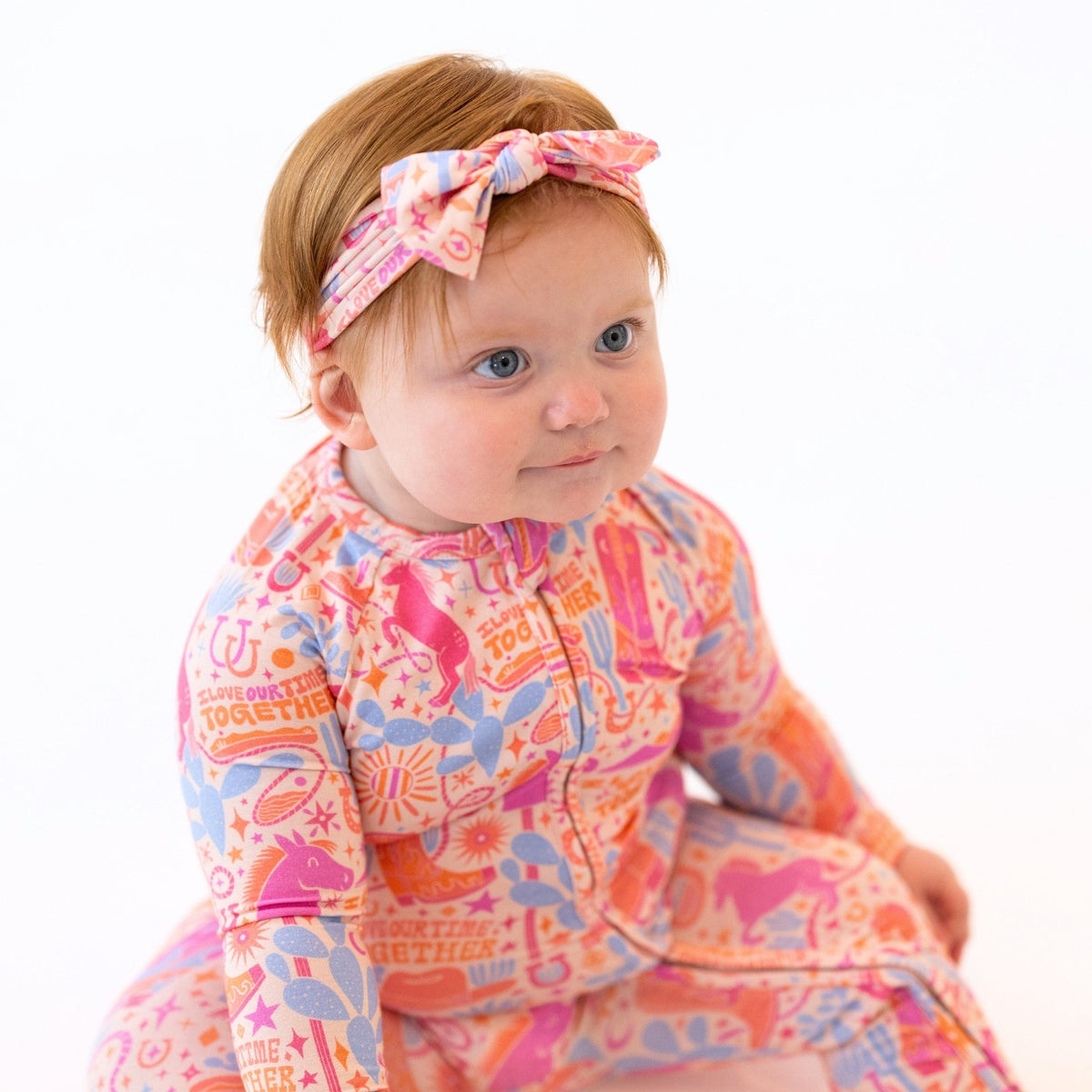 Baby in The Second-Skin Convertible Romper Saddle Up with pink-blue print and a headband.