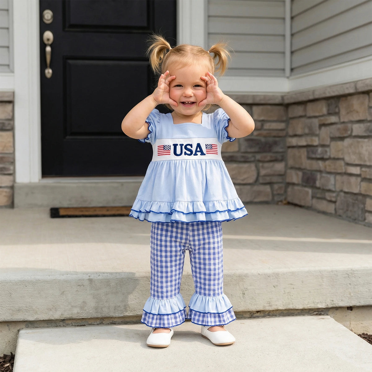 Child wearing a blue outfit with 'USA' text and American flag designs, standing on a porch.