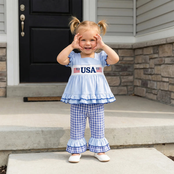 Child wearing a blue outfit with 'USA' text and American flag designs, standing on a porch.