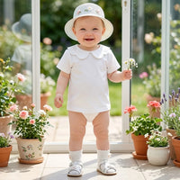 A baby standing in a bright sunroom wearing a white short-sleeve Peter Pan collar bodysuit, a white sun hat with small floral embroidery, white socks, and white sandals, while holding a small white flower. Potted flowers line the windows in the background.