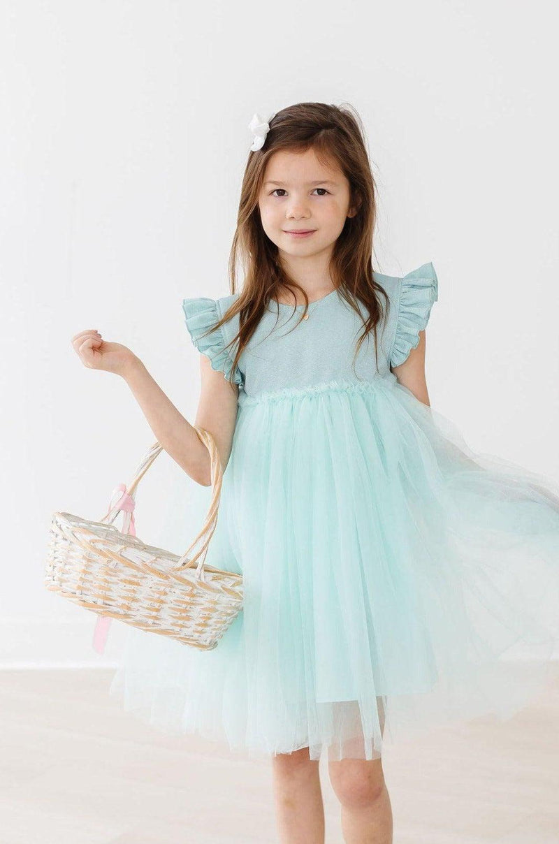 A young girl holding a basket, wearing a Sage Shimmer Tutu Dress from Mila & Rose, smiling at the camera in a bright, airy setting, perfect for showcasing her charming outfit.