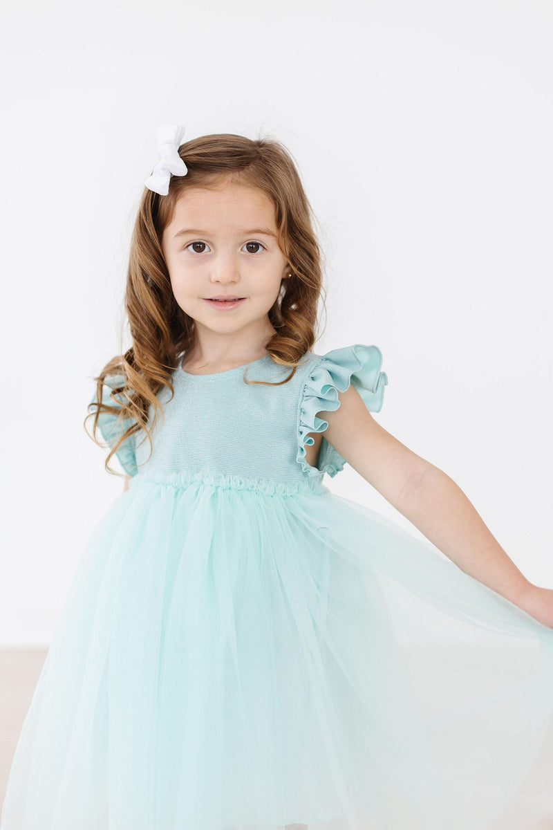 A close-up of a girl in the Sage Shimmer Tutu Dress from Mila & Rose, showcasing the flutter sleeves and delicate tulle skirt as she smiles confidently.
